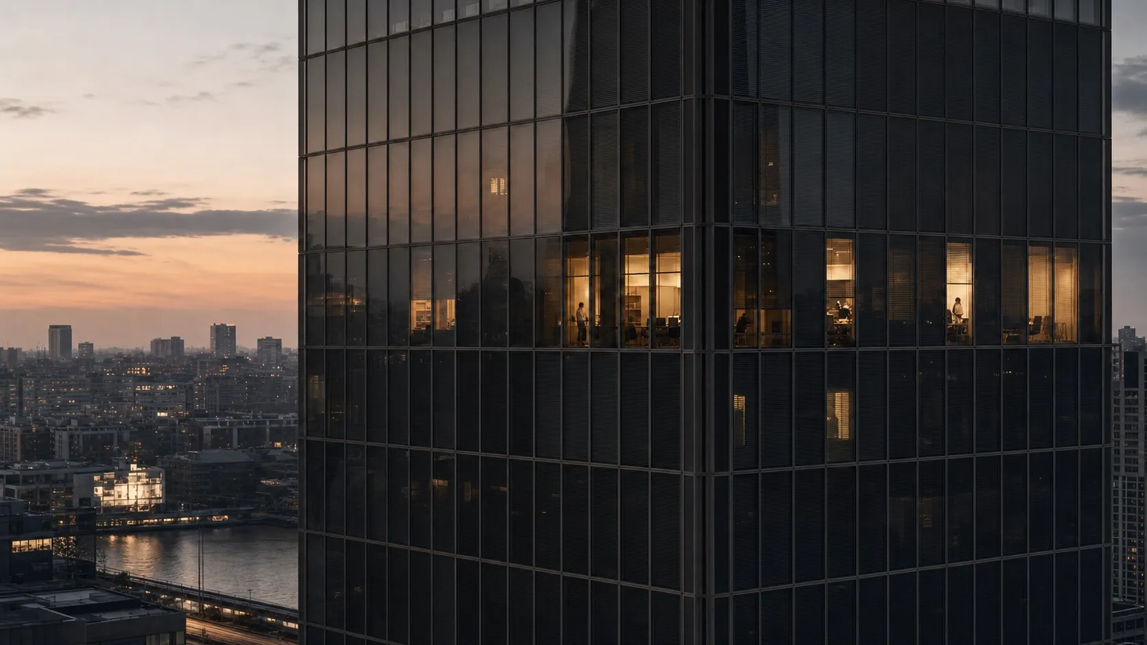 Ein gläsernes Hochhaus in der Abenddämmerung, in einigen Büros brennt Licht, dahinter Fluss und Stadt